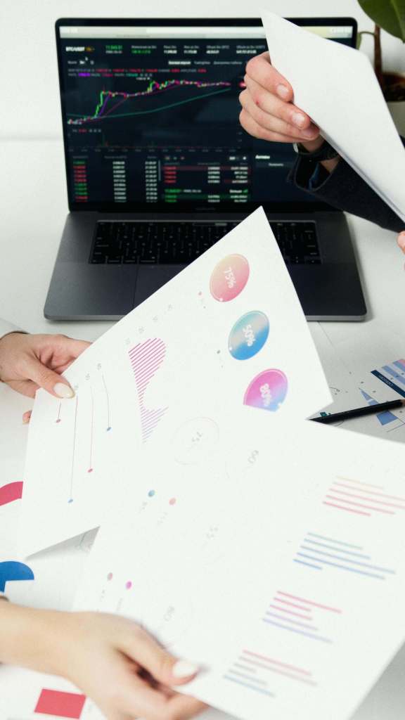 Два человека обсуждают документы с графиками и диаграммами, на столе лежит ноутбук с отображаемыми финансовыми данными.